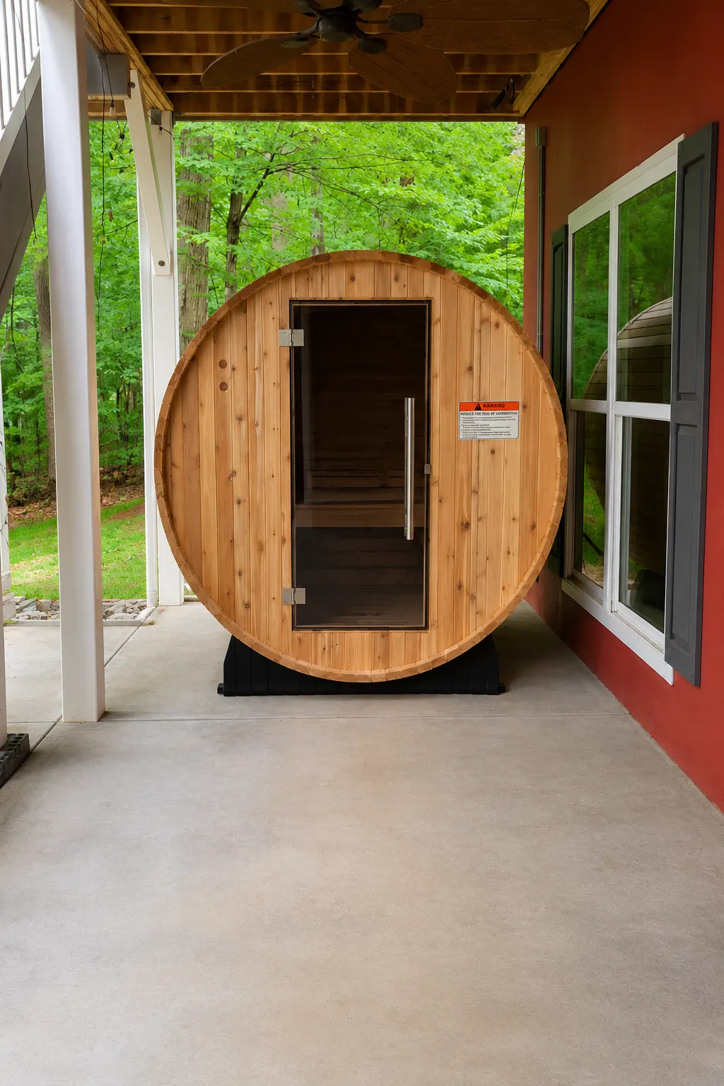 Cedar barrel sauna on the covered backyard patio of the red lake house, with a glass door and trees beyond